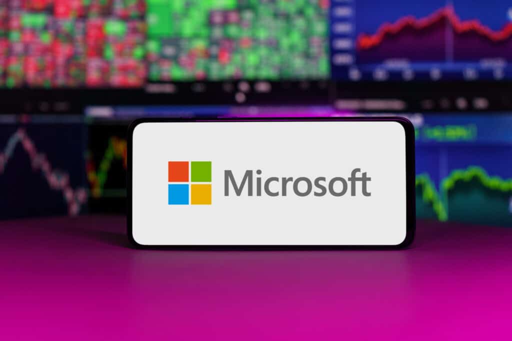 A smartphone displays the Microsoft logo on its screen, with colorful stock market charts and graphs blurred in the background.